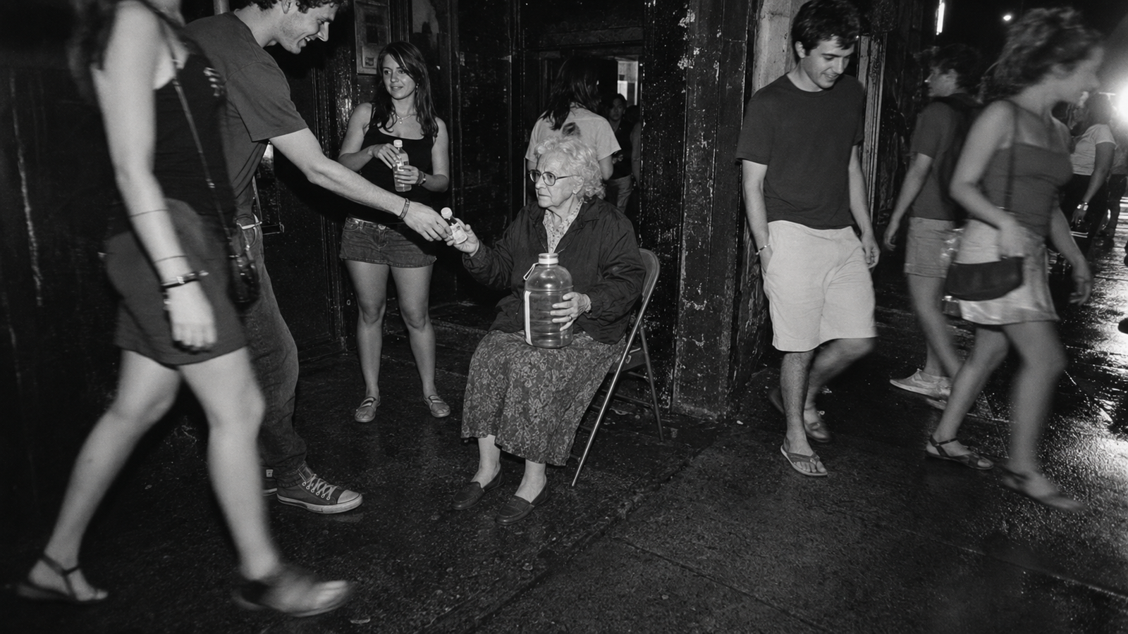 An elderly woman seated with a large container receiving a small bottle, in black and white.